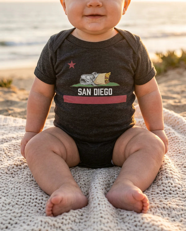 Baby wearing San Diego Burrito Flag bodysuit in black on a San Diego beach at golden hour