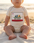 Baby wearing San Diego Burrito Flag bodysuit in white on a San Diego beach at golden hour