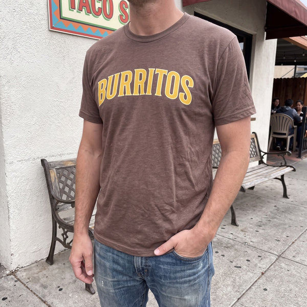 Man wearing a heather brown Burritos varsity T-shirt outside a taco shop with patio benches