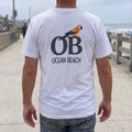 Man on a beach boardwalk in a white Ocean Beach T-shirt, back view showing a macaw perched on OB letters