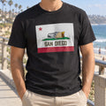 Man wearing a black T-shirt with a rectangle San Diego burrito flag print on a beach boardwalk with palm trees